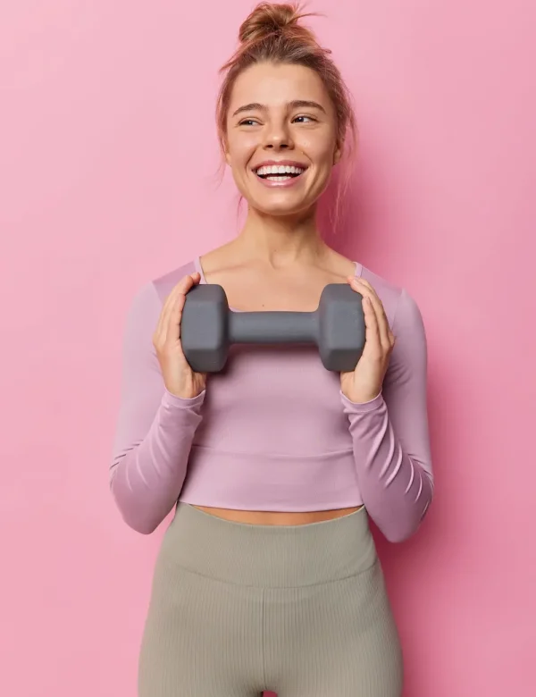 vertical-shot-healthy-fit-sportswoman-has-regular-training-gym-holds-dummbell-smiles-gladfully-leads-active-lifestyle-dressed-tracksuit-isolated-pink-background-people-sport-scaled